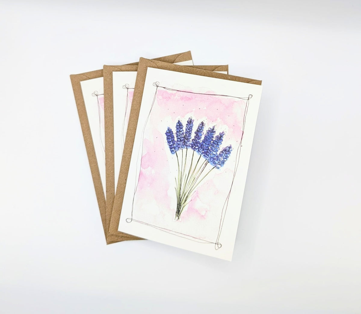 Hand-painted watercolour greeting card featuring a bunch of lavender stems on a blush pink background with an ink border, shown with kraft envelopes