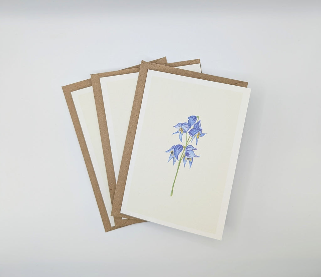 Hand-painted watercolour greeting card featuring a delicate blue delphinium stem with yellow centres on a white background, shown with kraft envelopes