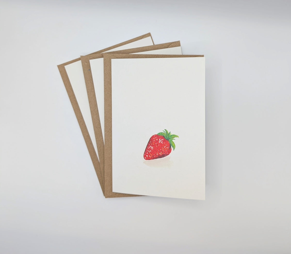 Hand-painted watercolour greeting card featuring a single plump strawberry, shown with kraft envelopes
