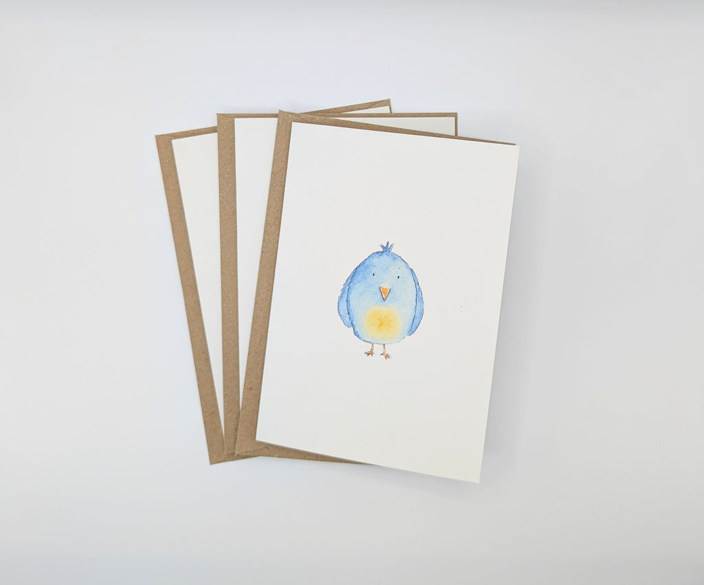 Hand-painted watercolour greeting card featuring a round fluffy blue bird with an orange beak and yellow chest, shown with kraft envelopes
