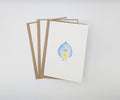 Hand-painted watercolour greeting card featuring a round fluffy blue bird with an orange beak and yellow chest, shown with kraft envelopes