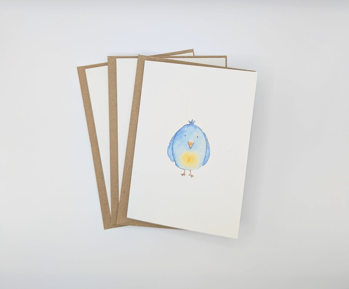 Hand-painted watercolour greeting card featuring a round fluffy blue bird with an orange beak and yellow chest, shown with kraft envelopes