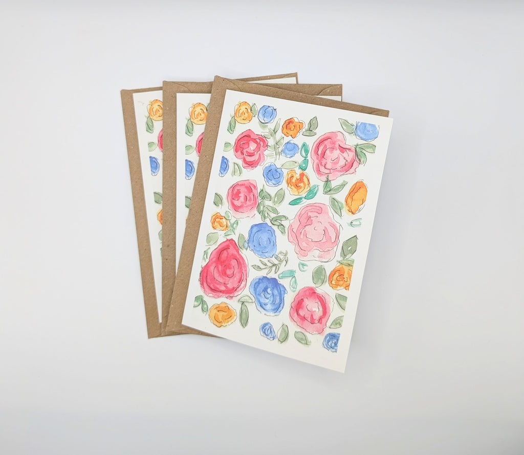 Hand-painted watercolour greeting card covered in pink, blue and orange roses with green leaves, shown with kraft envelopes