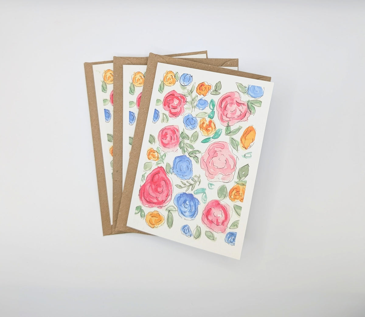 Hand-painted watercolour greeting card covered in pink, blue and orange roses with green leaves, shown with kraft envelopes