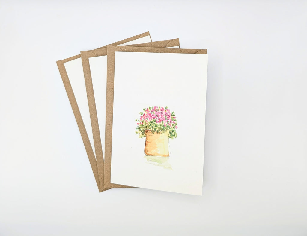 Hand-painted watercolour greeting card featuring a terracotta pot overflowing with tiny pink flowers and green foliage, shown with kraft envelopes