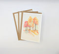 Hand-painted watercolour greeting card featuring fiery autumn trees in red, orange and gold lining a winding path, shown with kraft envelopes