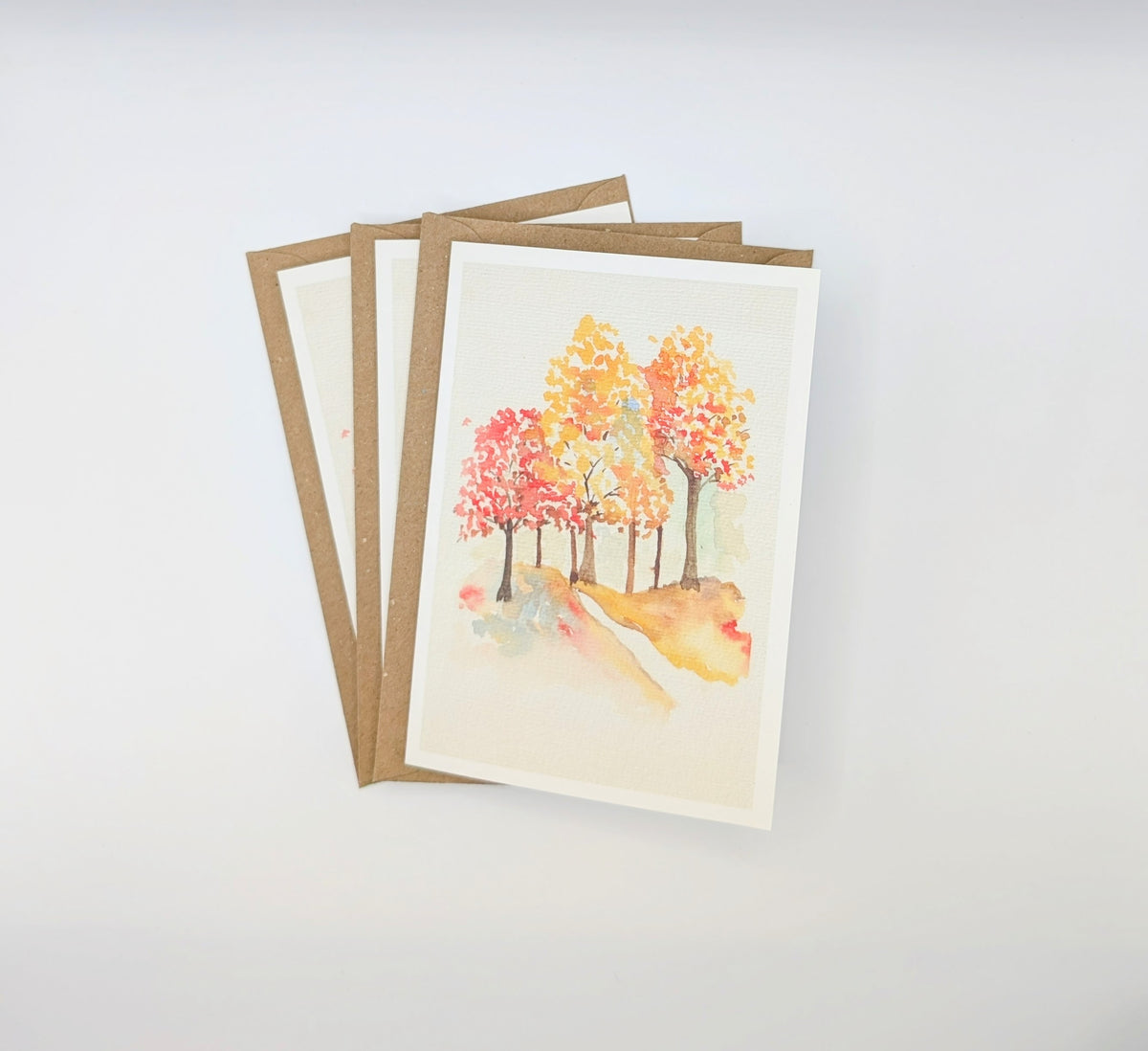 Hand-painted watercolour greeting card featuring fiery autumn trees in red, orange and gold lining a winding path, shown with kraft envelopes